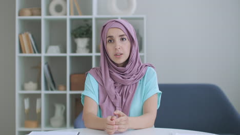 A-Muslim-woman-in-a-hijab-sits-at-a-table-and-looks-into-the-camera-waving-her-hand-and-talking.-vlog-or-video-call-a-woman-in-a-hijab.-shot-of-the-head-of-an-Arab-woman-in-conversation